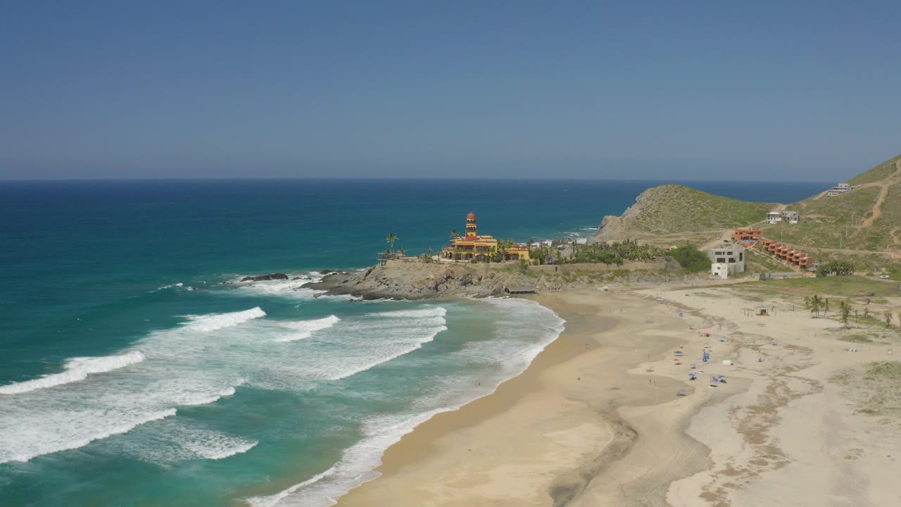 Aerial view over Cerritos, Baja California Sur, with white surf bands entering from the left, a broad sandy beach at right, a rocky point with low buildings, and no visible people.