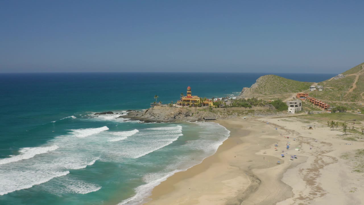 Aerial view in Cerritos, Baja California Sur, with turquoise water in the foreground, broken white waves near shore, hillside roads at right, coastal structures, and no visible people.