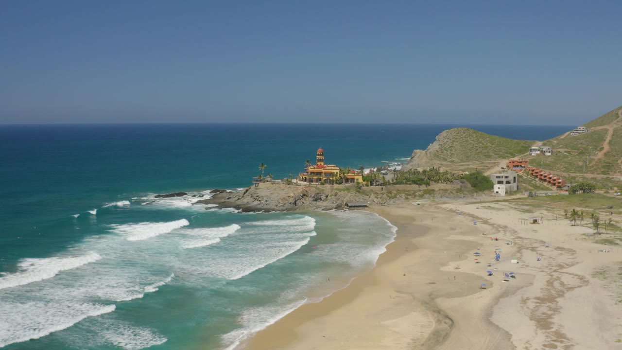 Drone view of surf, beach sand, rocky coastline, low buildings, palms, and open horizon.