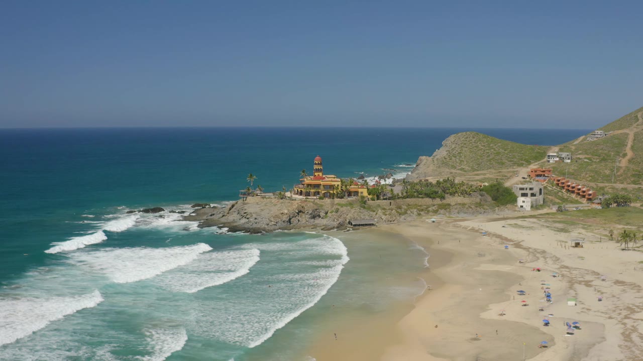 Aerial view in Cerritos, Baja California Sur, where a broad white wave band moves closer to the beach below a rocky point, yellow building, hillside, and no visible people.