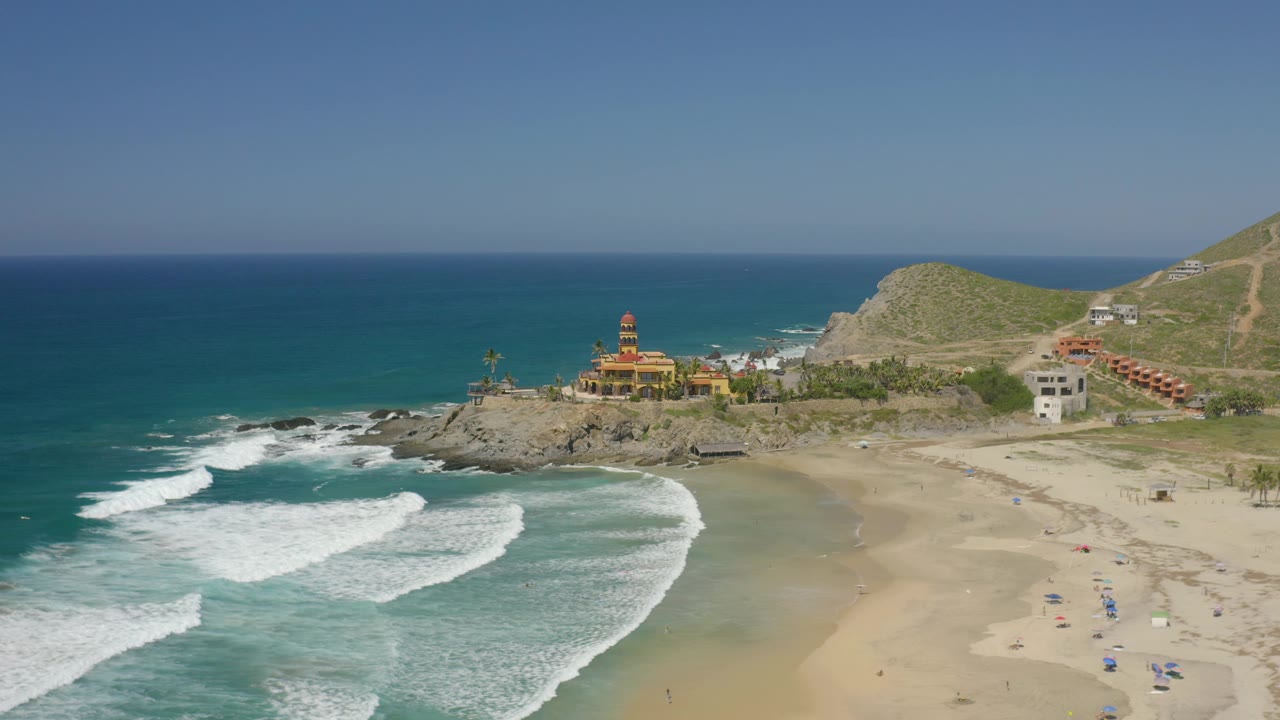Cerritos, Baja California Sur beach from above, with layered surf lines at left, a sandy cove at right, hillside buildings, a yellow structure on the rocks, and no visible people.