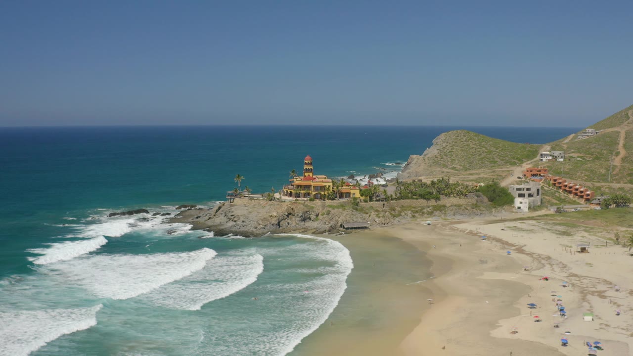 Mid-clip aerial frame over Cerritos, Baja California Sur, with larger white foam patches in the foreground, open tan sand, the rocky point, coastal buildings, and no visible people.