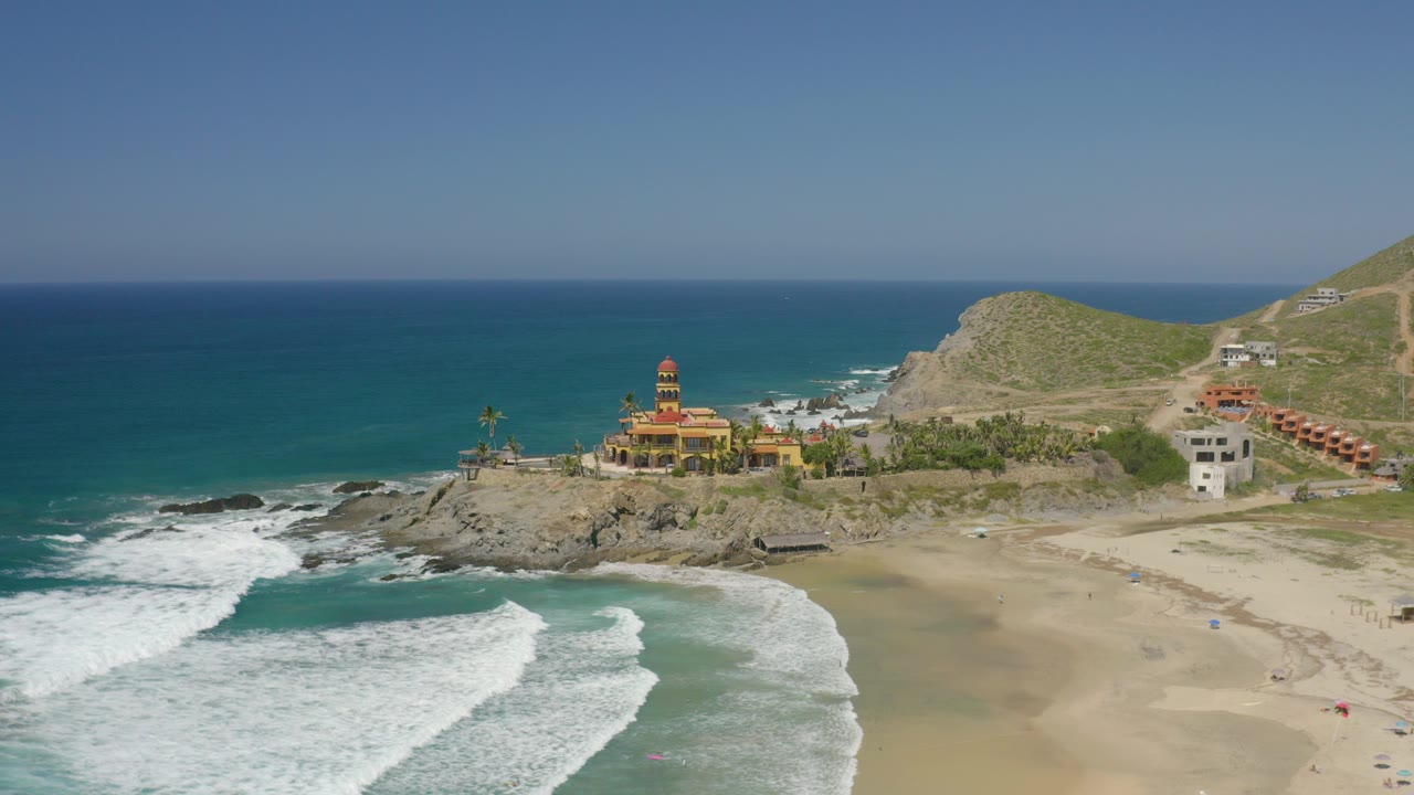 Aerial frame over Cerritos, Baja California Sur showing white surf entering from the lower left, open sand to the right, a rocky point with a yellow towered building, and no visible people.