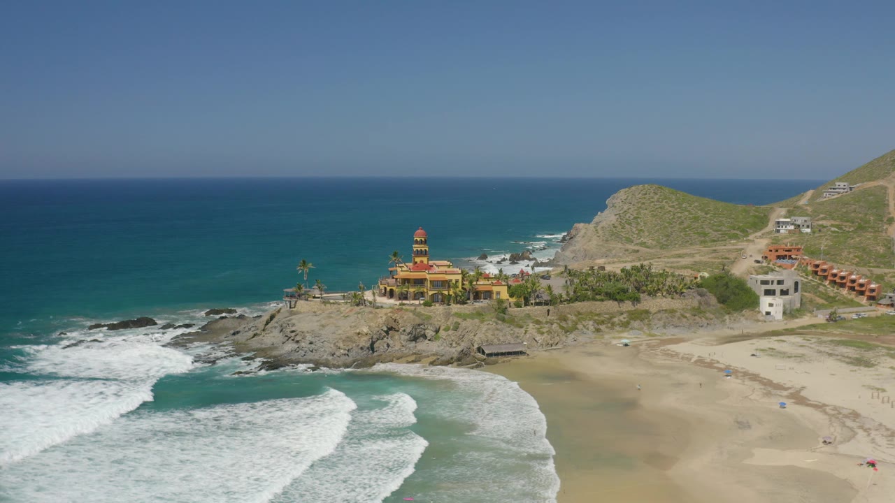 Wide drone frame over Cerritos with turquoise water across the left, whitewash reaching the lower beach, a rocky headland and towered yellow building near the center, and no visible people.