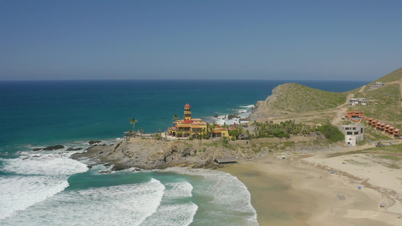 Aerial coastline with empty beach, surf, palm trees, and towered yellow building