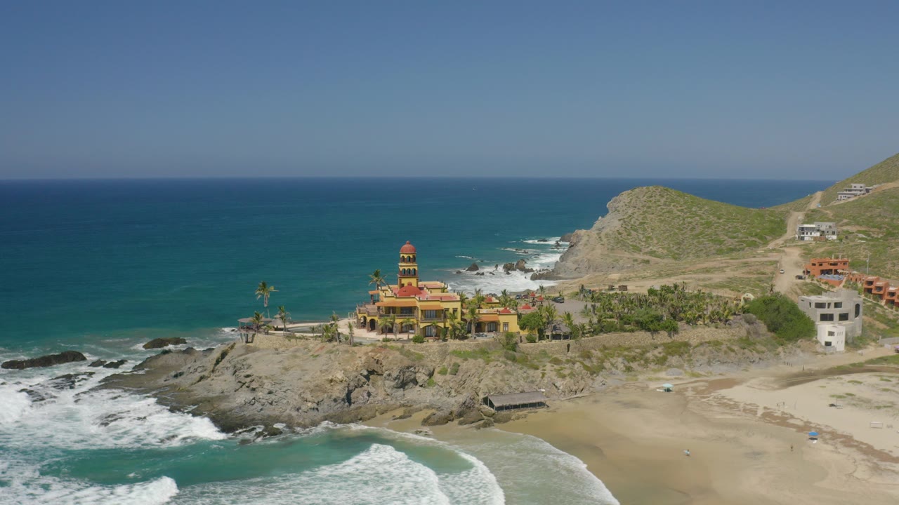 Aerial frame in Cerritos, Baja California Sur showing a yellow cliffside structure, rocky shoreline, turquoise water, sandy beach, dry hills, and no visible people.