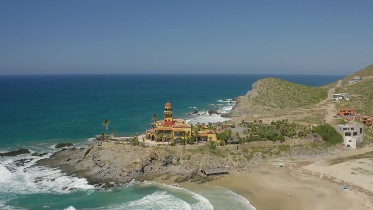 Aerial view in Cerritos showing the red-roofed structure on a rocky point, foam washing over shoreline rocks, beach sand at lower right, hillside buildings, and no visible people.