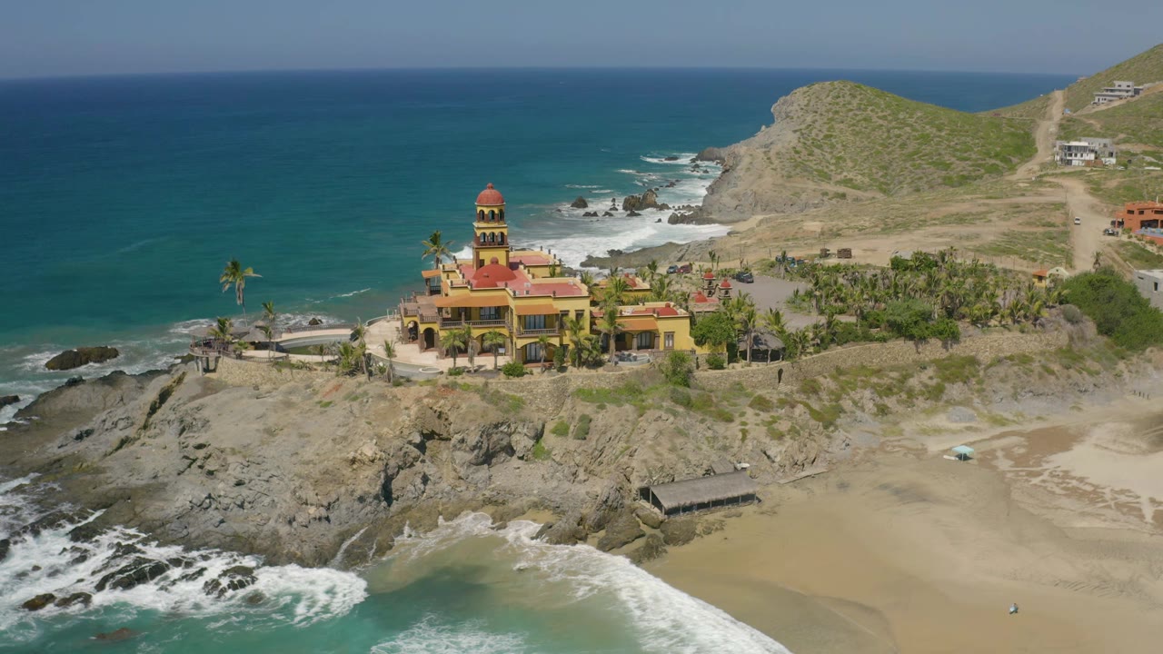 High aerial angle over a red-roofed coastal building, rocks, beach, and blue ocean.