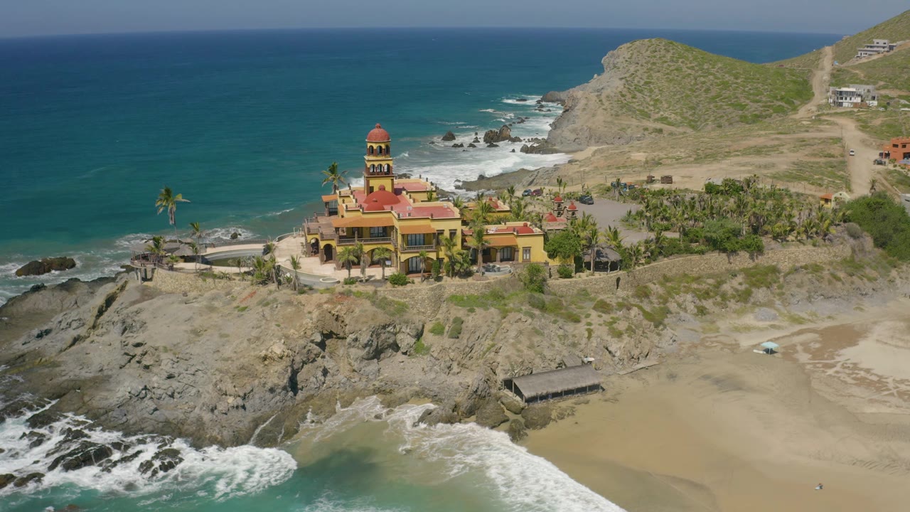 Drone-like view in Cerritos showing the towered yellow building, palms, curved shoreline rocks, blue water, dry hills, and no visible people in the beach area.