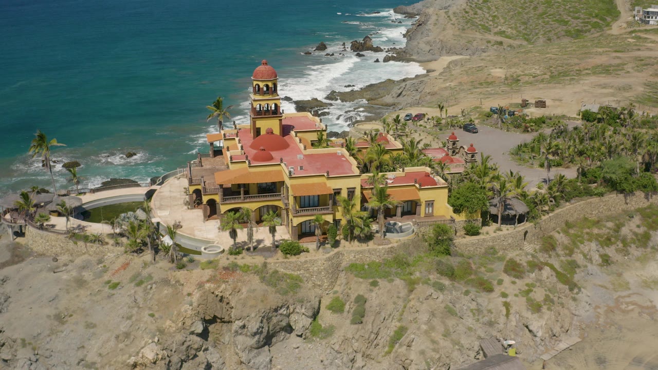 Aerial view in Cerritos, Baja California Sur of a yellow mansion with red roofs, a domed tower, palms, rocky shoreline, turquoise water, and no visible people.