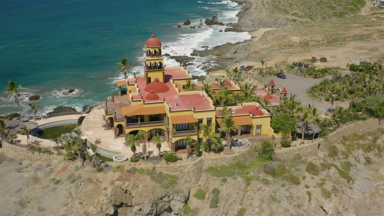 A closer aerial angle shows the Cerritos mansion's red roof planes, central domed tower, palm courtyard, rocky coast, and no visible people.