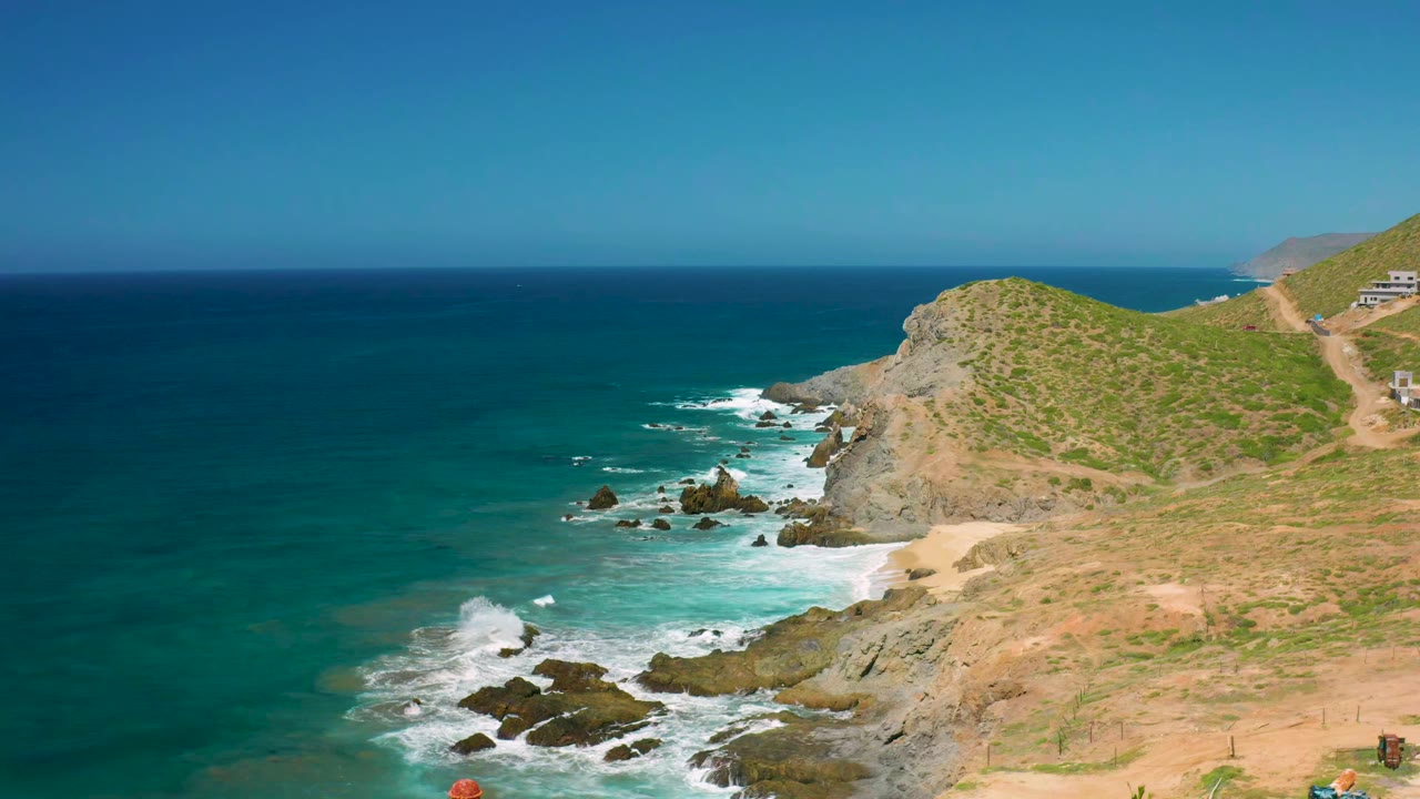 Cerritos coastal frame with the rocky surf line centered below a green-tan hill, clear sky above the horizon, and no visible people.
