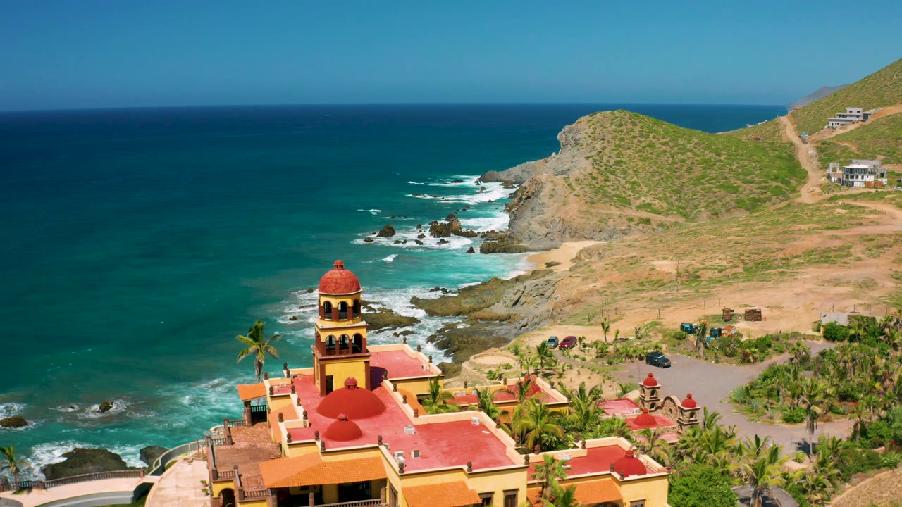 Cerritos aerial frame showing the red roof and tower of the coastal building, palm landscaping, a curving access area, blue water, white wave lines, and dry hillside terrain.