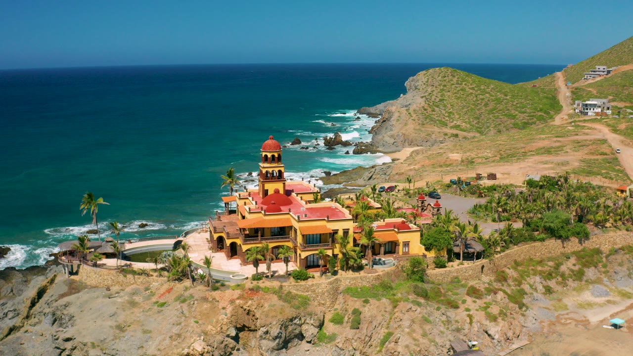 Aerial view in Cerritos, Baja California Sur, showing a red-domed yellow building among palms, rocky shoreline, blue ocean, dry hills, and no visible people at the start of the clip.