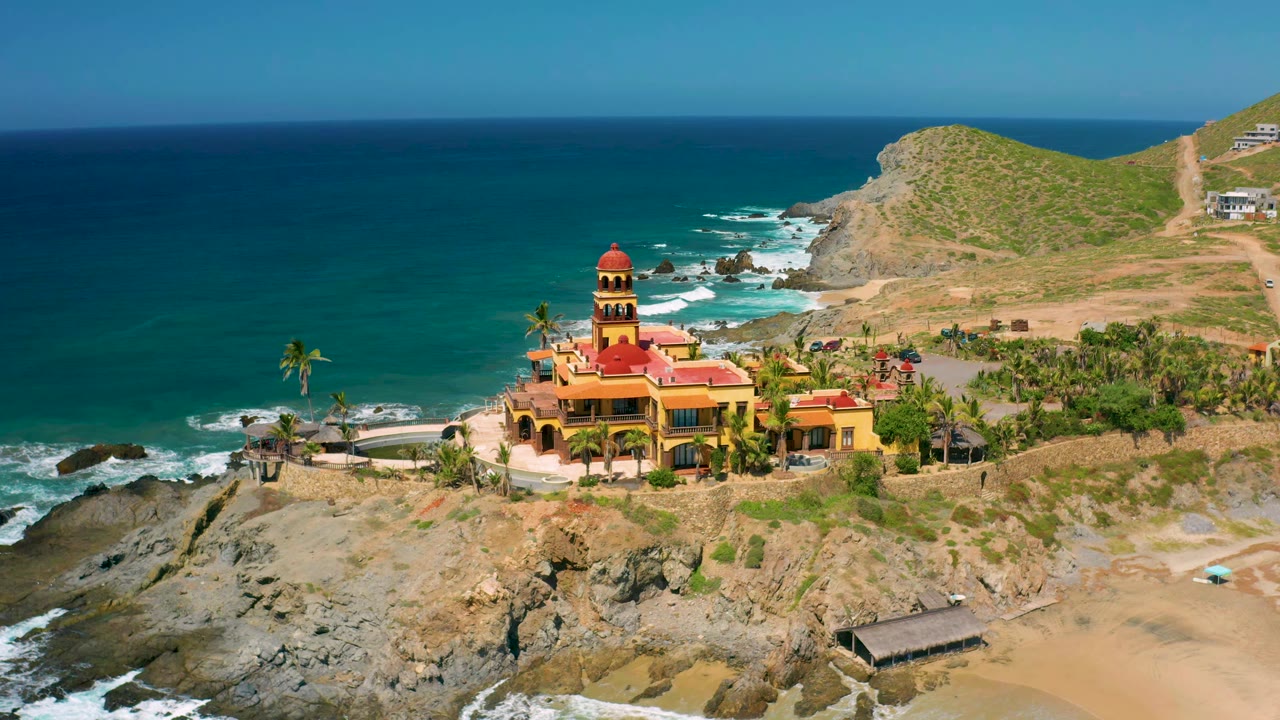 At 0:01, the aerial view keeps the Cerritos building centered while white surf curls around the rocks below and the hillside fills the right background, with no visible people.