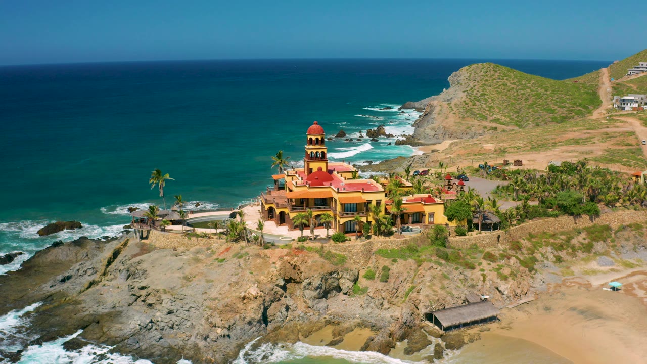 At 0:02, the Cerritos shoreline view shows the red-roofed complex slightly right of center, with exposed rock ledges, foamy water, beach sand, and no visible people.