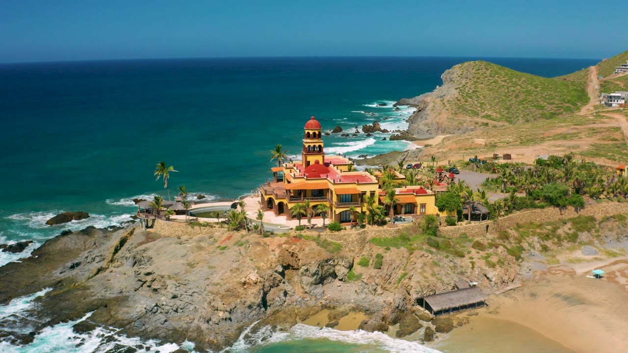 At 0:03, the drone angle places more open ocean to the left of the Cerritos building complex, with waves breaking in the foreground and no visible people on the beach or rocks.