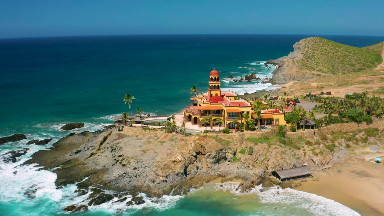 Cerritos aerial view with the building centered near the shoreline, white waves spreading across dark rocks, turquoise water at lower left, dry hill behind, and no visible people.