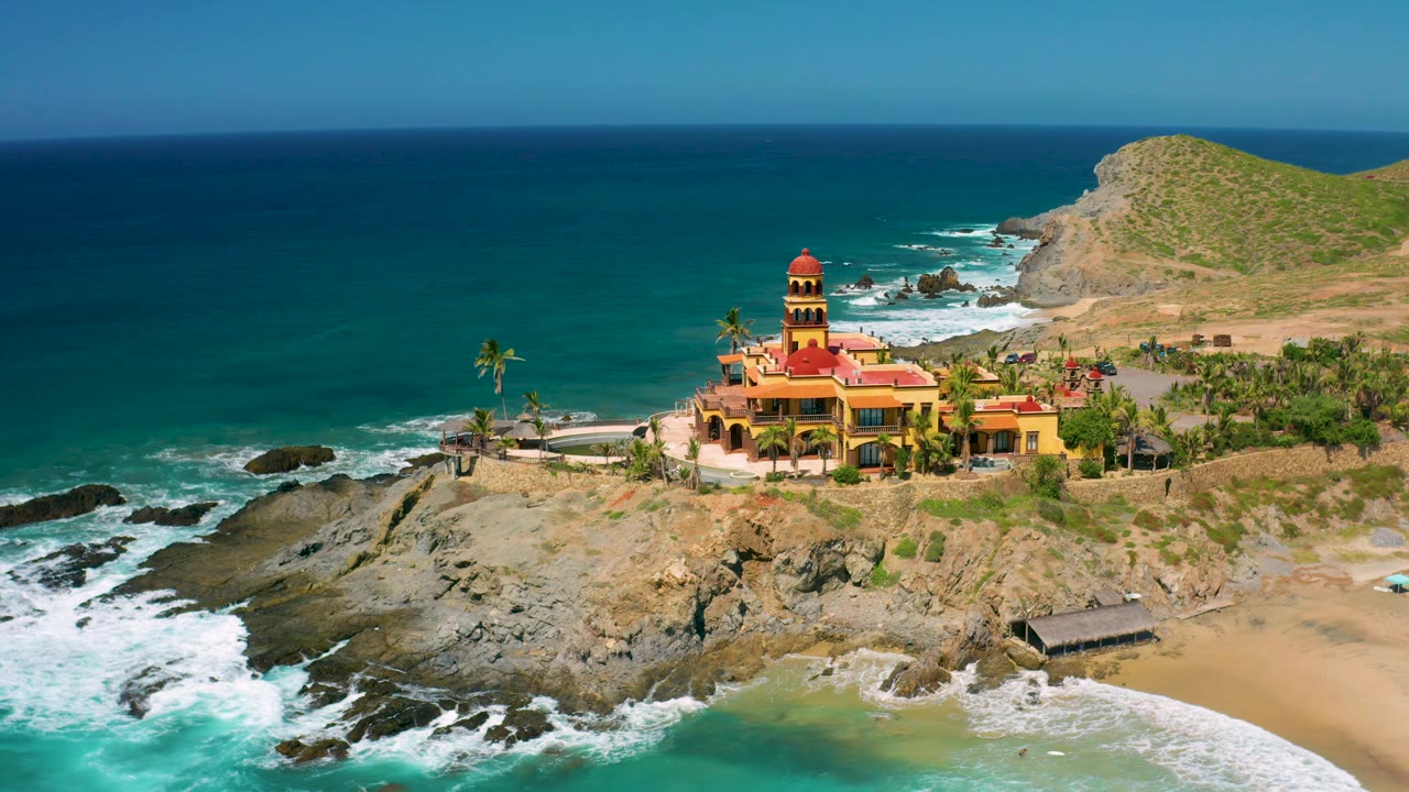 Aerial frame in Cerritos, Baja California Sur with the red-roofed building slightly right of center, foamy water around the rocks, beach sand at lower right, ocean horizon, and no visible people.