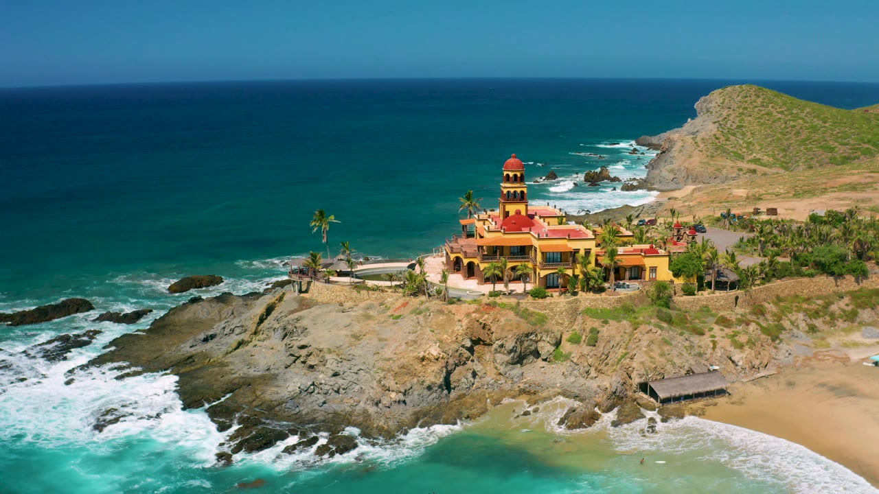 Cerritos coast frame with a broad blue ocean background, traditional building on the rocky point, white surf pushing into the foreground rocks, sandy beach at right, and no visible people.