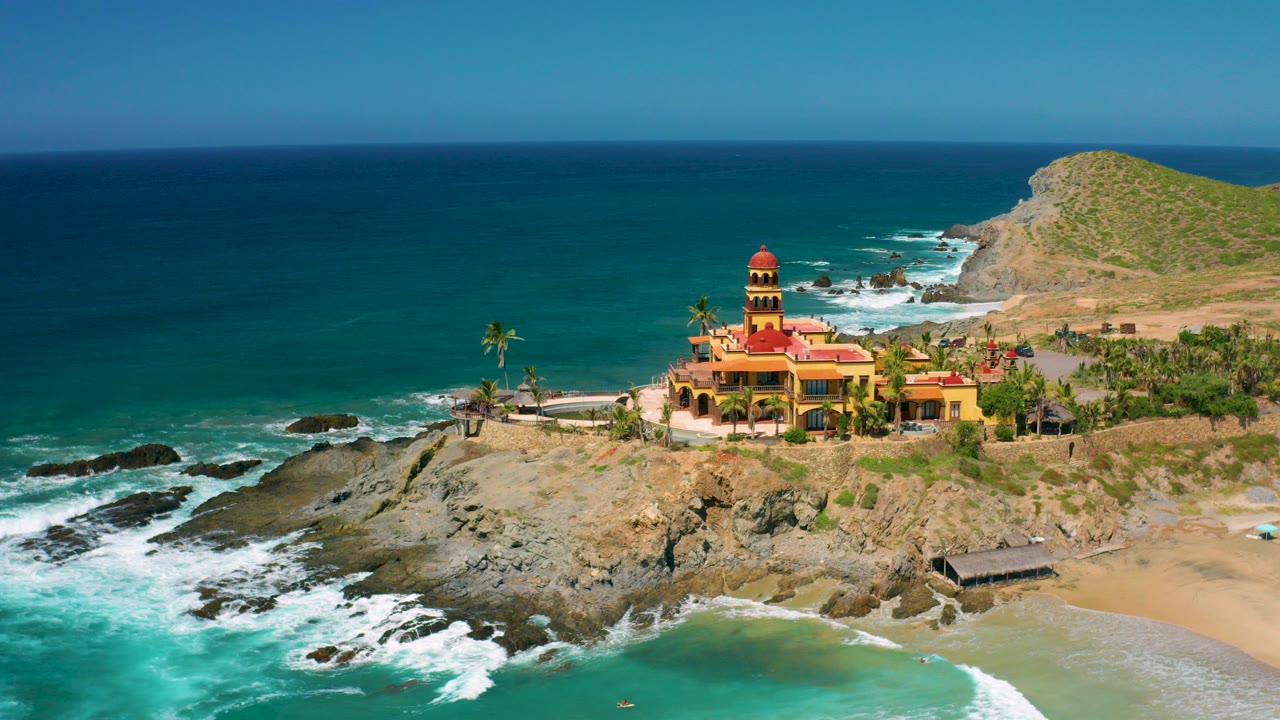 At 0:00, an aerial view over Cerritos, Baja California Sur shows teal ocean, white surf on rocks, sandy beach, palms, a yellow coastal building with red roofs, and no visible people.