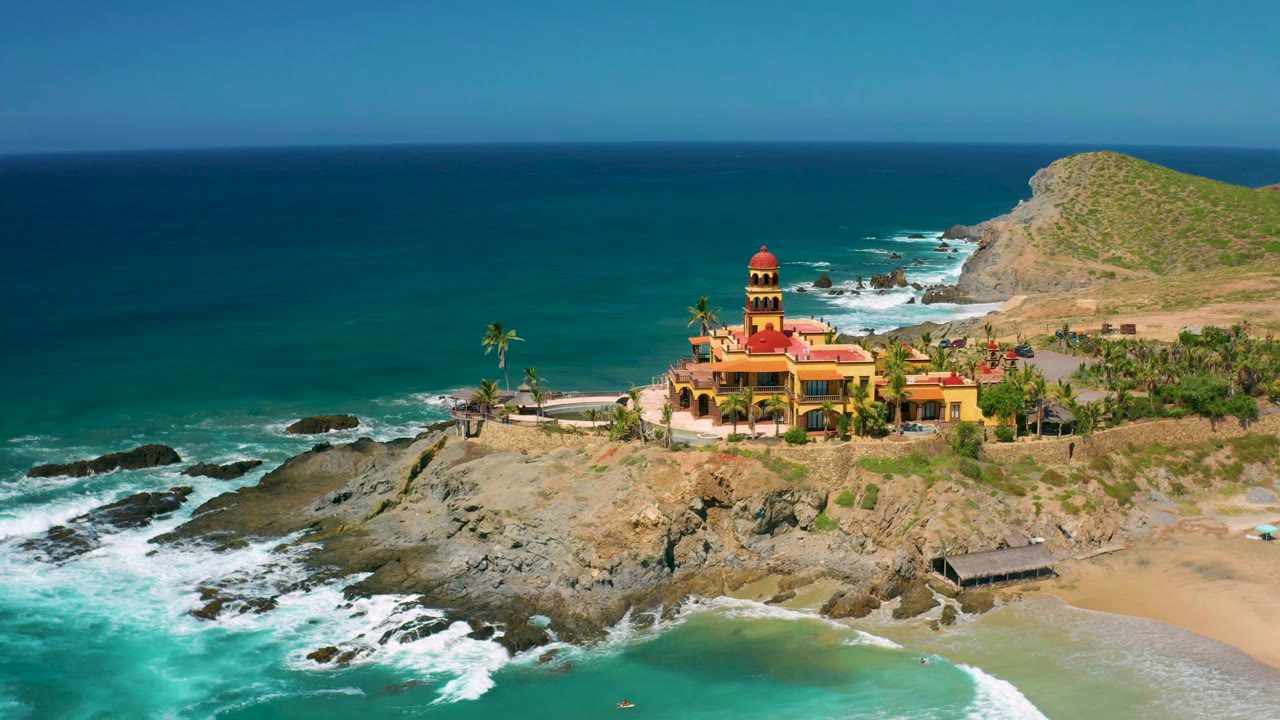 At 0:01, the drone view keeps the Cerritos shoreline centered, with whitewater spreading farther across the lower-left rocks, the red-roofed building above the beach, and no visible people.