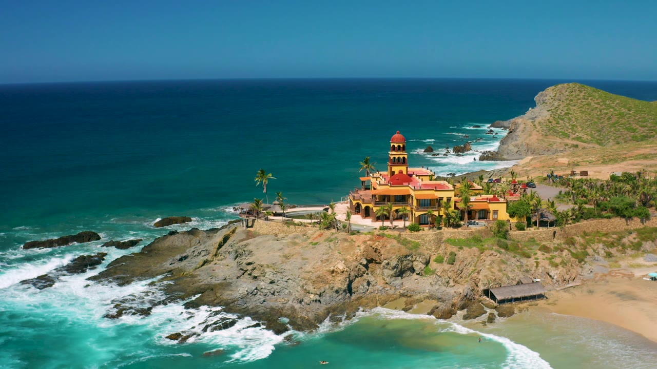 At 0:02, Cerritos appears from a slightly wide aerial composition with the ocean occupying the left side, a sandy beach at lower right, the towered red-roof building near center, and no visible people.