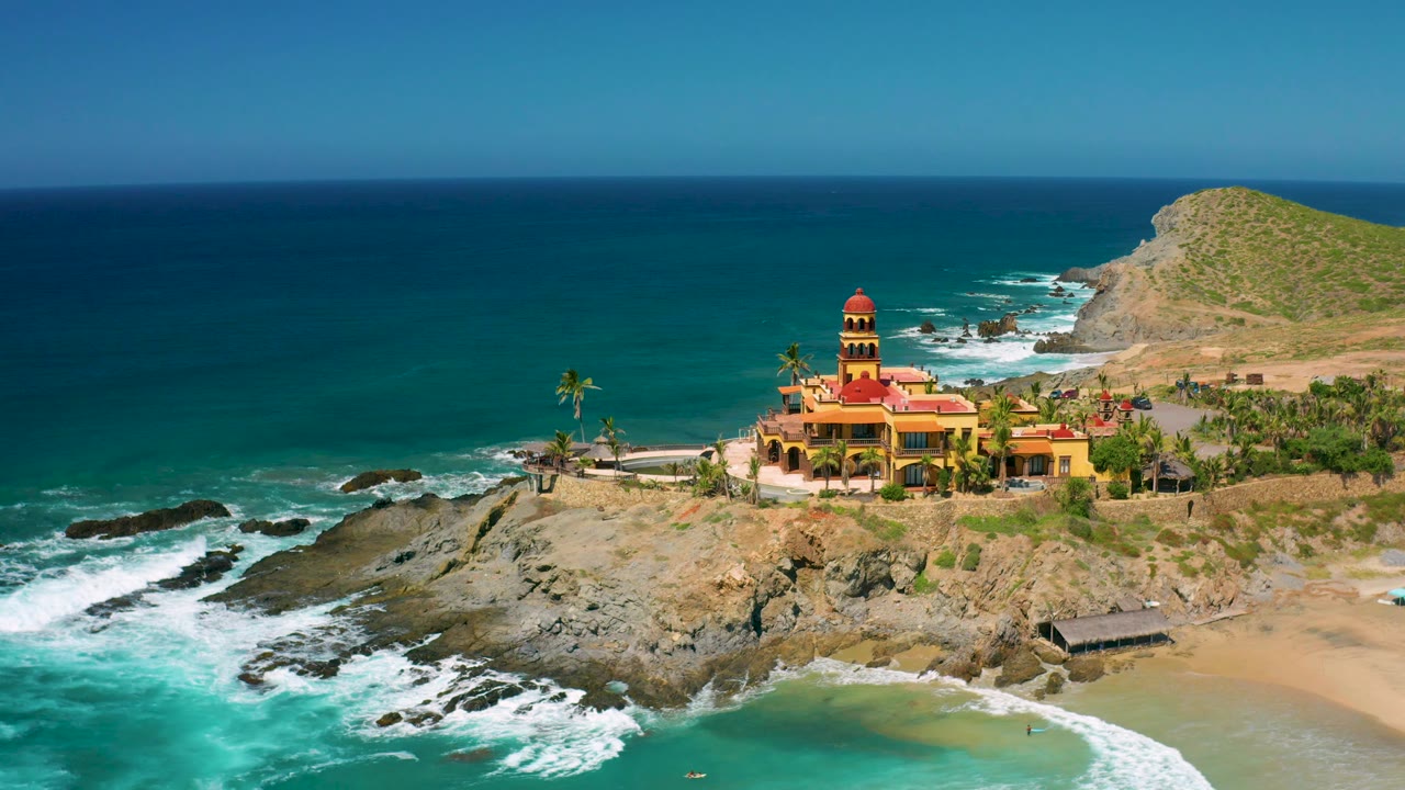 Drone-like view over surf and rocks toward a red-roofed building, palms, beach, and hillside.