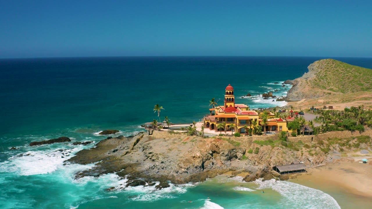Opening aerial frame over Cerritos, Baja California Sur, showing a yellow coastal building with red roofs, rocky shore, turquoise water, sandy beach, hillside, and no visible people.