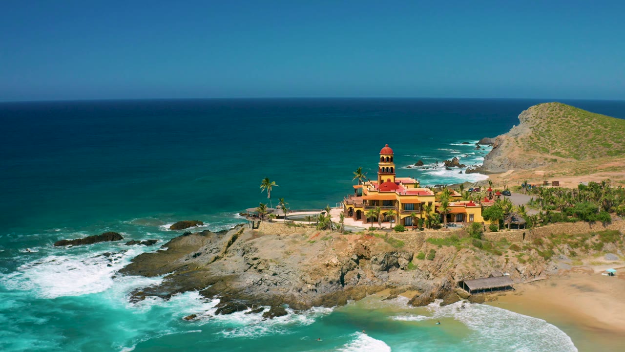 Cerritos coastline frame with the yellow building slightly right of center, palm trees around it, blue-green water to the left, pale beach to the right, and no visible people.