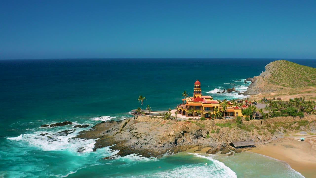 Aerial view in Cerritos, Baja California Sur of a red domed building on a rocky point with turquoise surf, tan beach, low hill, and no visible people.