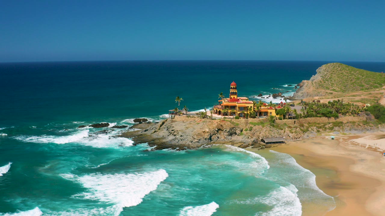Opening aerial view over Cerritos, Baja California Sur, with turquoise surf in the foreground, a rocky point and colorful beachfront building beyond it, and no visible people.