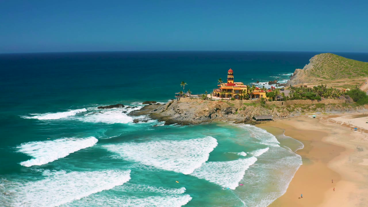 At one second, the Cerritos coastline remains wide in frame as white wave bands spread across shallow turquoise water below the rocky point, with no visible people.