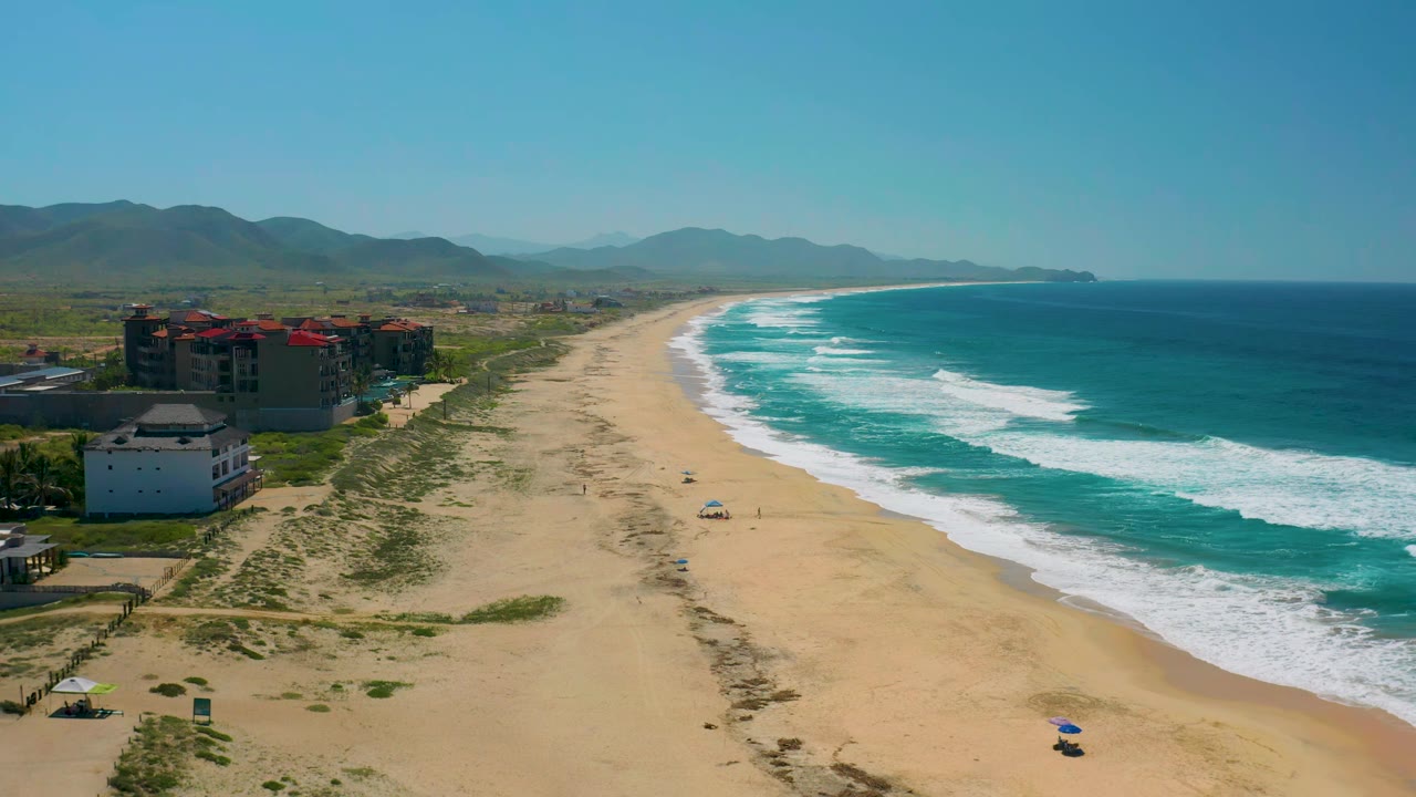 Wide aerial coastline with waves breaking beside sandy beach and buildings.