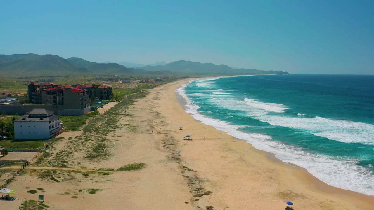 Cerritos aerial view with more open water on the right, a long pale beach through the center, scattered dune plants, modern coastal buildings at left, hazy mountains inland, and no visible people.
