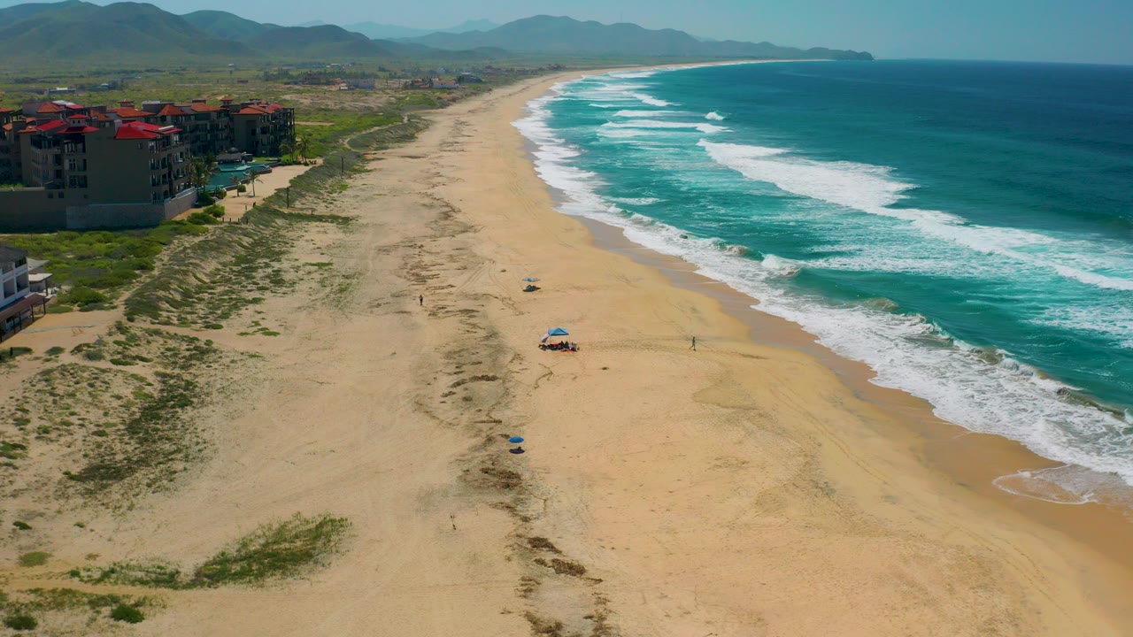 Aerial view over Cerritos, Baja California Sur, with red-roofed beachfront buildings at left, a wide sand beach below, turquoise surf at right, and tiny people visible on the shore.