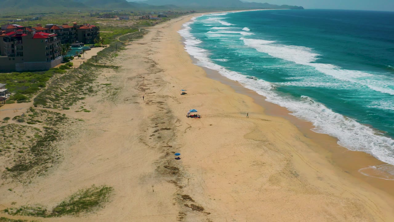 Cerritos beach from above, with red-roofed buildings close to the left edge, empty sand stretching into the distance, white surf bands on the right, and tiny beachgoers visible near umbrellas.
