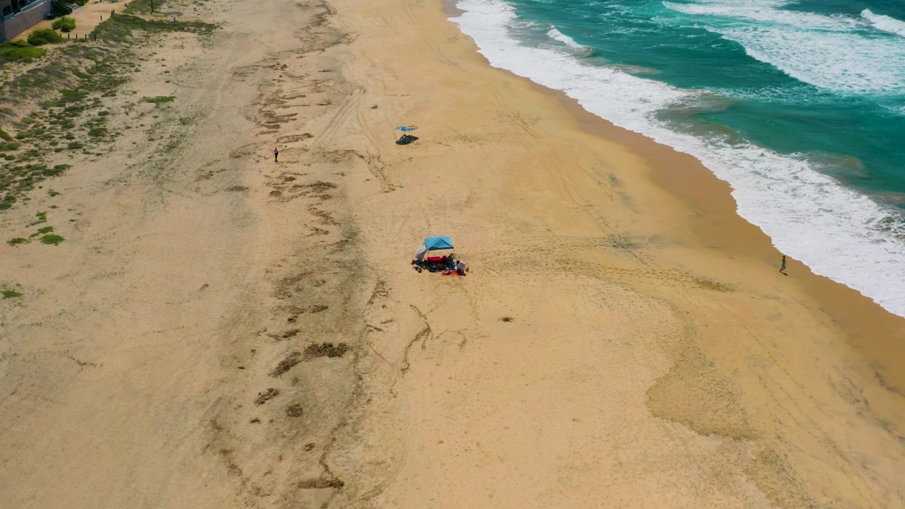 High aerial view over Cerritos beach in Baja California Sur, Mexico, with tan sand, surf along the right edge, dune vegetation to the left, a blue shade tent, and tiny distant people visible.