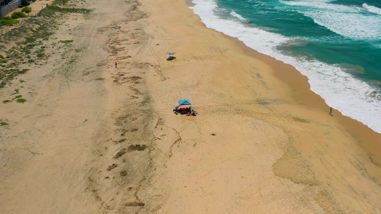 Aerial frame of Cerritos beach showing the surfline on the right, open tan sand through the center, dune scrub at the left edge, a blue shade tent near the lower frame, and tiny distant people visible.