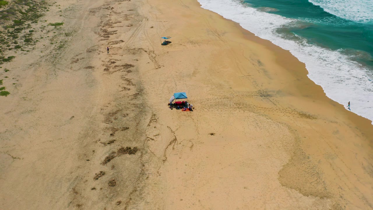 Wide overhead view at Cerritos beach with diagonal surf on the right, tire-marked sand and vegetation on the left, a blue shade tent below center, and tiny distant people visible near the beach setup and shoreline.