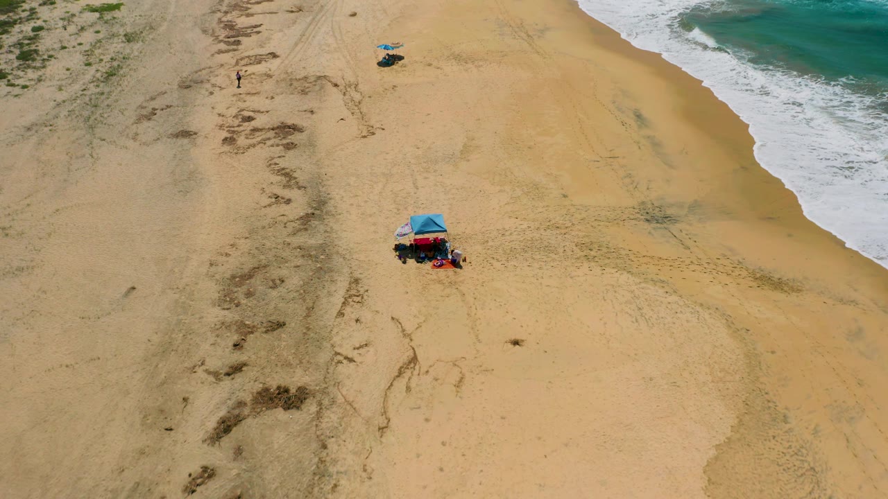 Aerial beach frame with blue tent, open sand, and surf on the right.