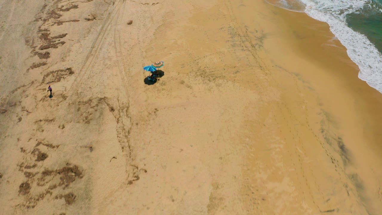 High aerial view over Cerritos beach sand with a blue umbrella setup near center, surf on the right edge, and tiny distant beachgoers visible on the left.