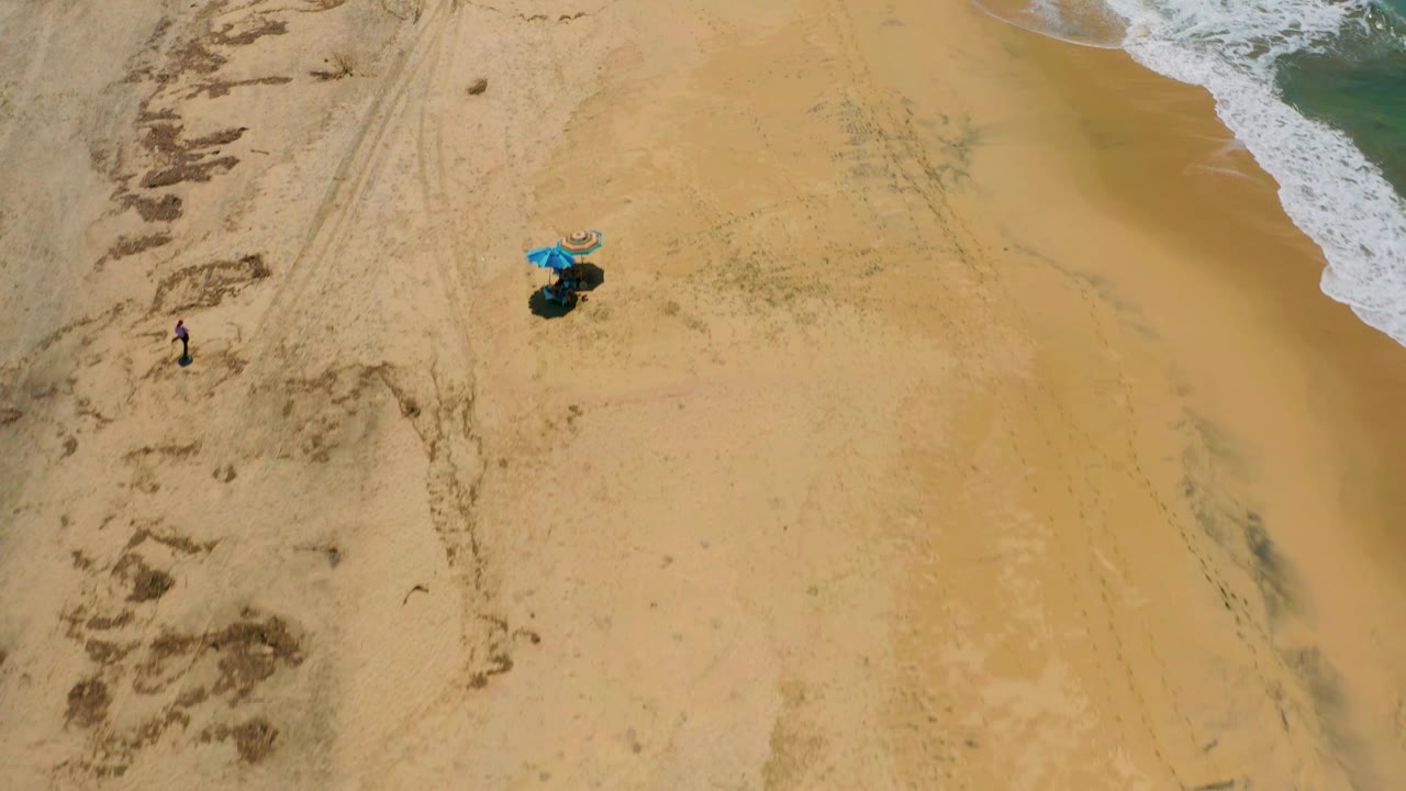 Aerial frame over Cerritos sand with the umbrella setup slightly right of center, white water at the upper right, and small distant beachgoers on the left side.