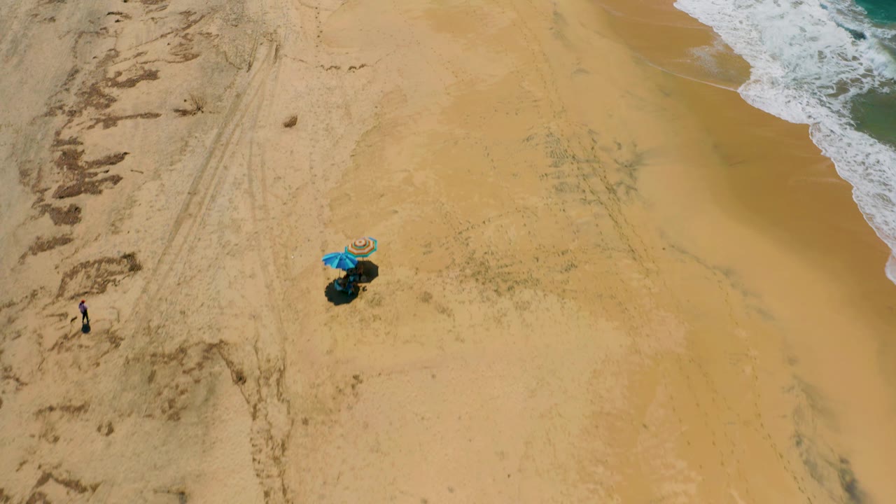 High drone-style view of Cerritos beach with a small umbrella setup near the lower middle, a tiny beachgoer to the left, and foamy surf entering from the upper right.