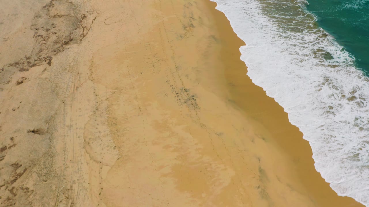 Diagonal shoreline with tan sand, foamy wave edge, and blue water.