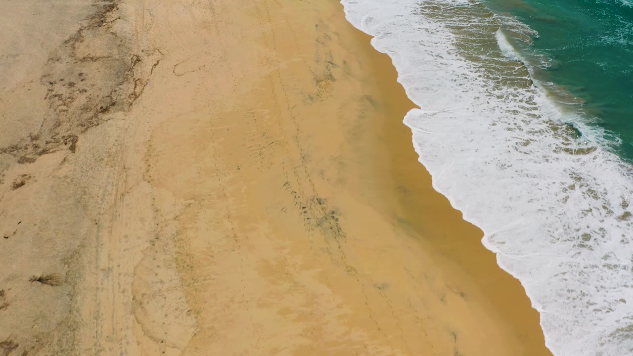Overhead Cerritos shoreline frame with the wave foam shifted farther along the right edge and broad empty sand on the left, no visible people.