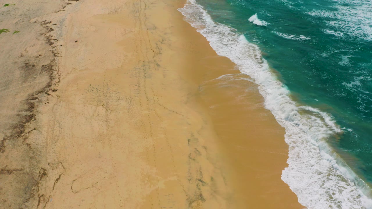 Top-down view of Cerritos beach with tan sand on the left, blue-green ocean on the right, white foam along the shore, and no visible people.