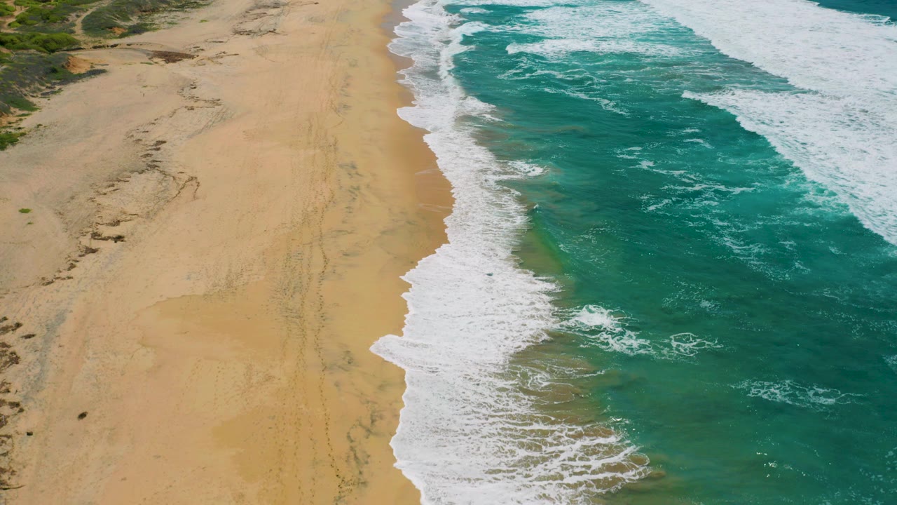 Aerial view over Cerritos, Baja California Sur with tan sand on the left, turquoise surf on the right, white foam along the shore, dune vegetation at the top edge, and no visible people.