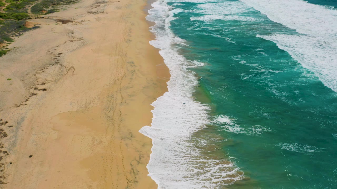 Aerial frame of Cerritos beach showing a diagonal line of white surf, broad tan sand, darker blue water offshore, faint tracks in the sand, and no visible people.