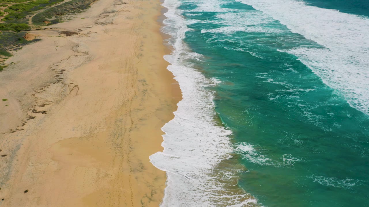 Aerial view over Cerritos, Baja California Sur with green-blue shallow water, white breaker lines, uninterrupted sand, low scrub near the upper beach, and no visible people.